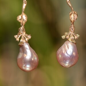 Puede incluir: Un par de pendientes de oro con una perla barroca rosa. Los pendientes tienen un delicado diseño floral.