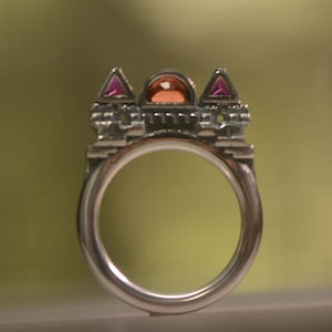 Puede incluir: Un anillo de plata con un diseño de castillo y piedras preciosas rosas. El anillo tiene una piedra redonda y transparente en el centro.