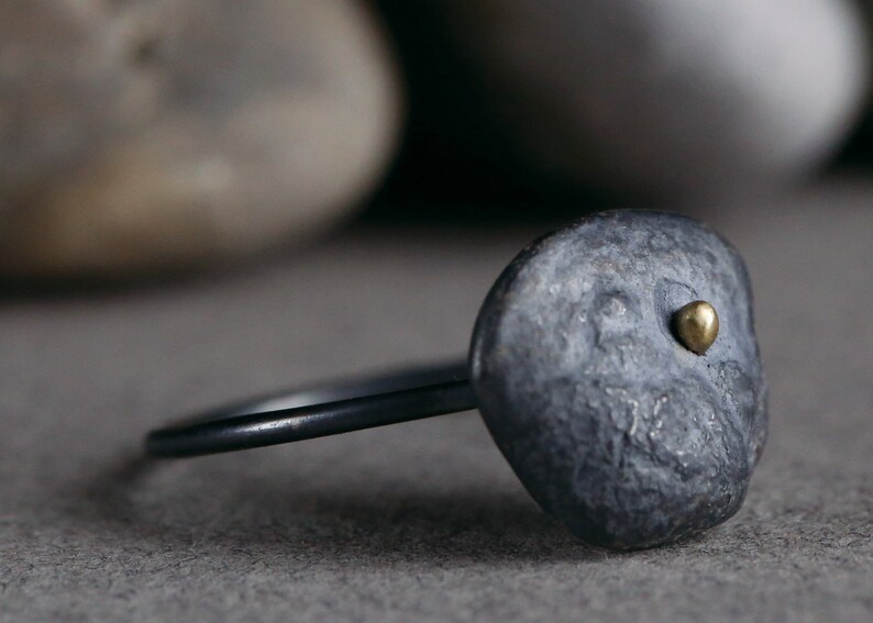 Puede incluir: Un anillo negro con una banda texturizada, gris, similar a una piedra y un peque&ntilde;o acento dorado. El anillo est&aacute; sobre una superficie gris.