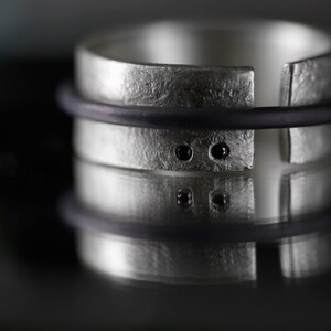 Op de afbeelding: Een zilveren ring met een getextureerde afwerking en twee zwarte edelstenen. De ring is aan &eacute;&eacute;n kant open en heeft een zwart elastiekje eromheen.