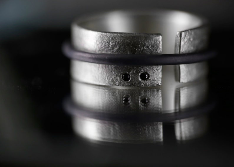 Op de afbeelding: Een zilveren ring met een getextureerde afwerking en twee zwarte edelstenen. De ring is aan &eacute;&eacute;n kant open en heeft een zwart elastiekje eromheen.