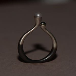 May include: A silver ring with a black and white pearl design. The ring is made of two curved bands that meet at the top, where the pearls are set.