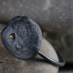 Puede incluir: Un anillo negro con una piedra gris texturizada y un peque&ntilde;o acento dorado. El anillo est&aacute; apoyado en una roca gris.