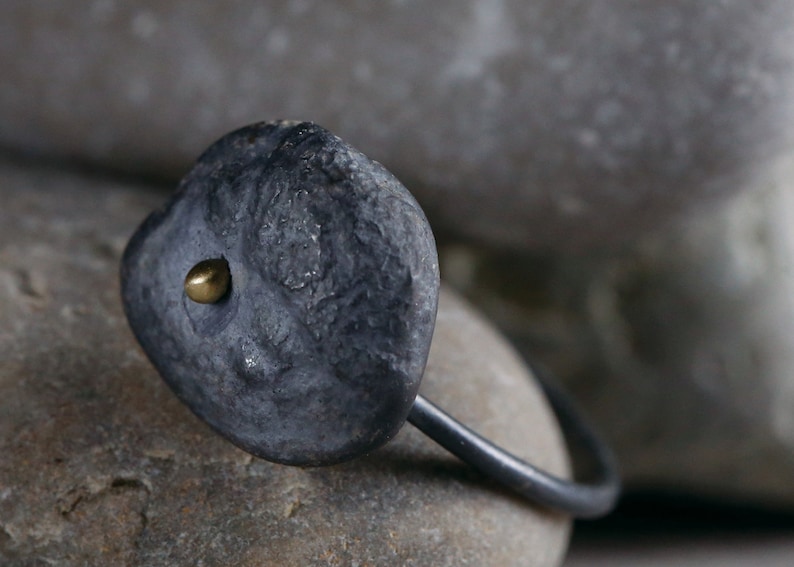 Puede incluir: Un anillo negro con una piedra gris texturizada y un peque&ntilde;o acento dorado. El anillo est&aacute; apoyado en una roca gris.