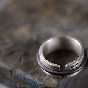 Op de afbeelding: Een zilveren ring met een opening in de band, en een zwarte rubberen band eromheen gewikkeld.