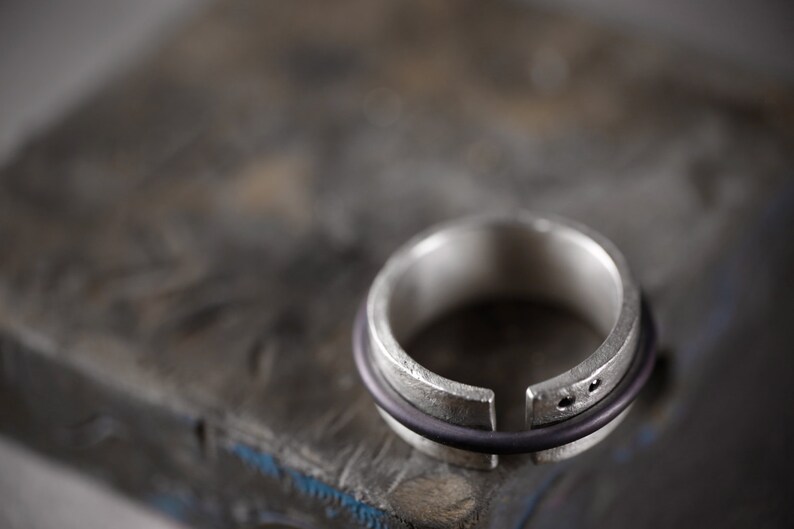 Op de afbeelding: Een zilveren ring met een opening in de band, en een zwarte rubberen band eromheen gewikkeld.