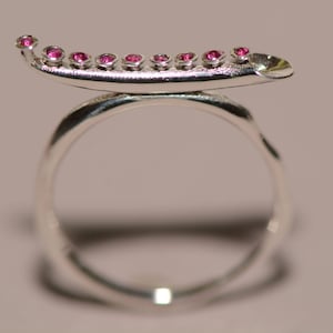 Peut inclure: Une bague en argent avec une bande incurvée et une rangée de pierres précieuses roses serties sur le dessus. La bague est conçue pour ressembler à un petit bateau.