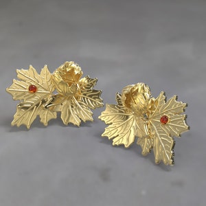 May include: A pair of gold-coloured leaf-shaped earrings. Each earring features a cluster of leaves with a small, round, orange-red gemstone in the centre. The earrings have a detailed, textured surface and a polished finish.