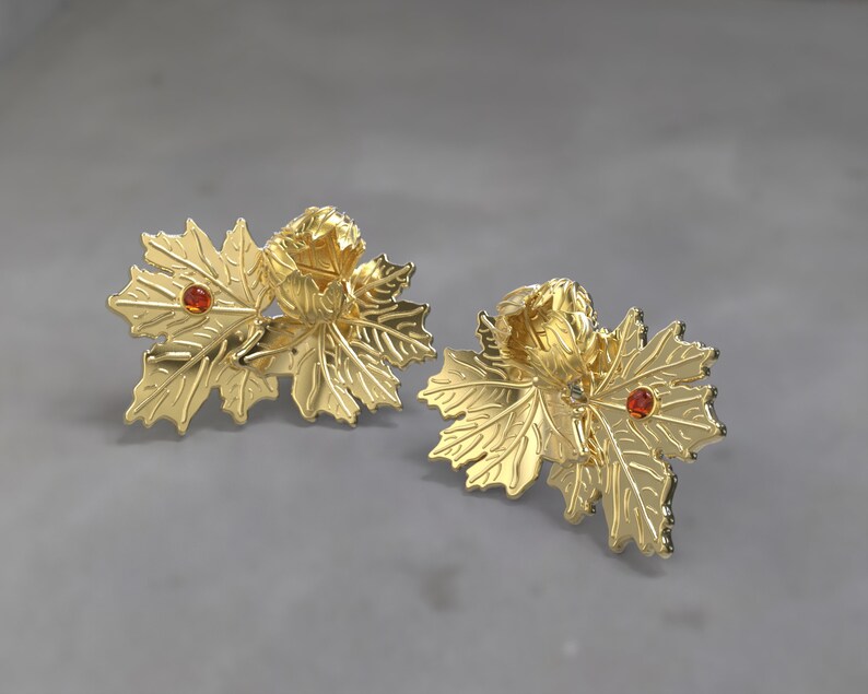 May include: A pair of gold-coloured leaf-shaped earrings. Each earring features a cluster of leaves with a small, round, orange-red gemstone in the centre. The earrings have a detailed, textured surface and a polished finish.