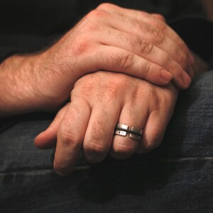 Op de afbeelding: Een close-up van iemands handen met een zilveren ring aan de ringvinger van de rechterhand. De ring heeft een getextureerd ontwerp en is gemaakt van twee banden.