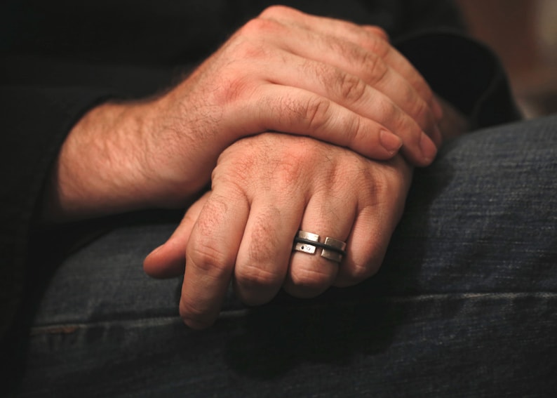 Op de afbeelding: Een close-up van iemands handen met een zilveren ring aan de ringvinger van de rechterhand. De ring heeft een getextureerd ontwerp en is gemaakt van twee banden.