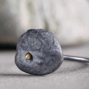 Puede incluir: Un anillo de plata con una piedra ovalada texturizada de color gris y un peque&ntilde;o punto dorado en el centro.