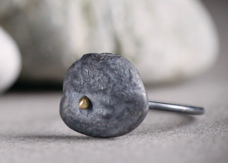 Puede incluir: Un anillo de plata con una piedra ovalada texturizada de color gris y un peque&ntilde;o punto dorado en el centro.