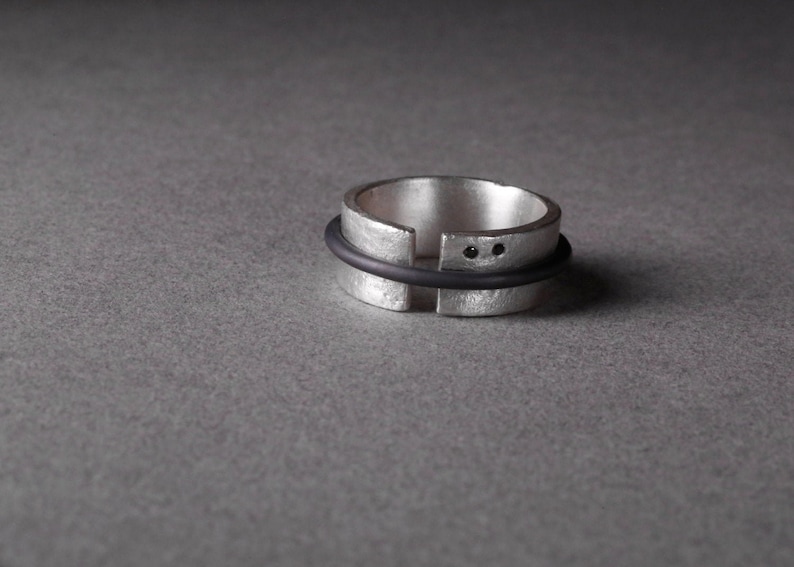 Op de afbeelding: Een zilveren ring met een zwart rubberen bandje eromheen. De ring heeft twee kleine zwarte stippen aan de bovenkant.