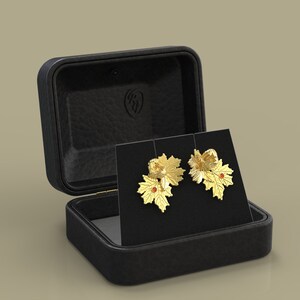 May include: Gold-coloured leaf earrings with red accents, presented on a black card within an open black leather jewellery box. The earrings have detailed leaf designs, suggesting a nature theme.
