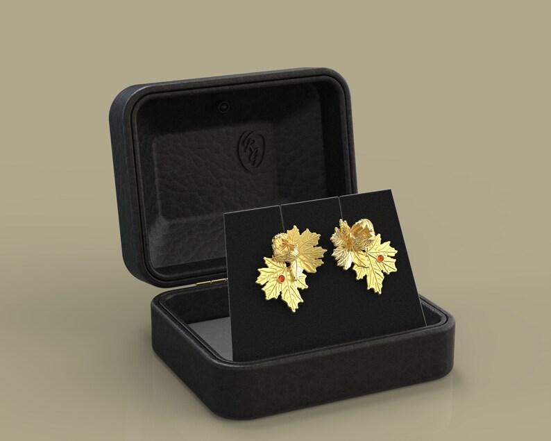 May include: Gold-coloured leaf earrings with red accents, presented on a black card within an open black leather jewellery box. The earrings have detailed leaf designs, suggesting a nature theme.