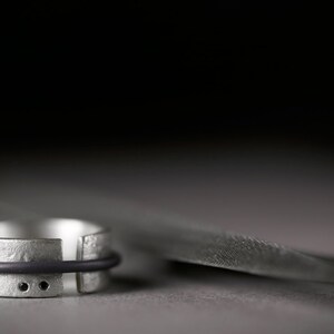 Op de afbeelding: Een zilveren ring met een zwart elastiekje en twee kleine zwarte stippen. De ring ligt op een grijs oppervlak.