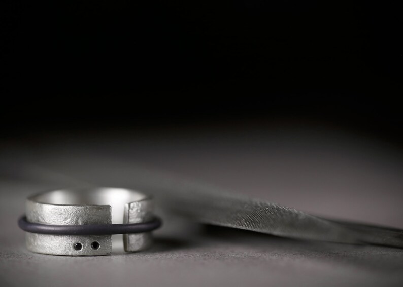 Op de afbeelding: Een zilveren ring met een zwart elastiekje en twee kleine zwarte stippen. De ring ligt op een grijs oppervlak.