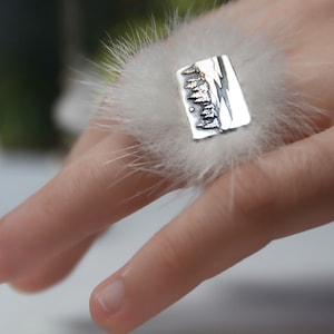May include: A silver ring with a textured design of a mountain range. The ring is on a finger wearing a white fluffy ring holder.
