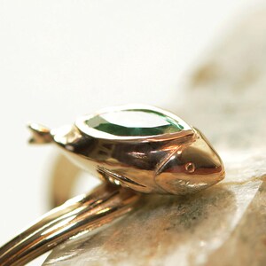 Puede incluir: Un anillo de oro con una piedra preciosa verde engastada en un dise&ntilde;o con forma de pez.
