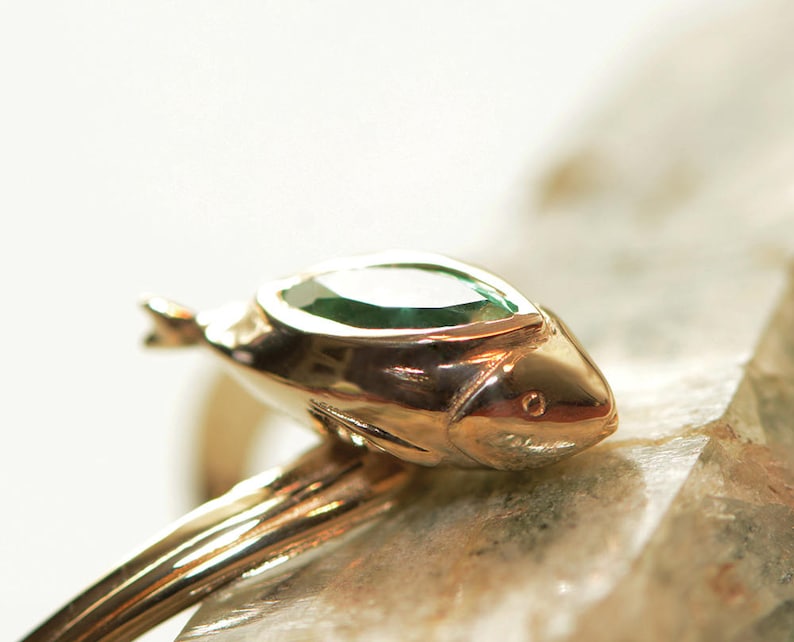 Puede incluir: Un anillo de oro con una piedra preciosa verde engastada en un dise&ntilde;o con forma de pez.