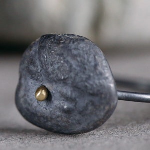 Puede incluir: Un anillo negro con una forma irregular texturizada y un peque&ntilde;o punto dorado en el centro.