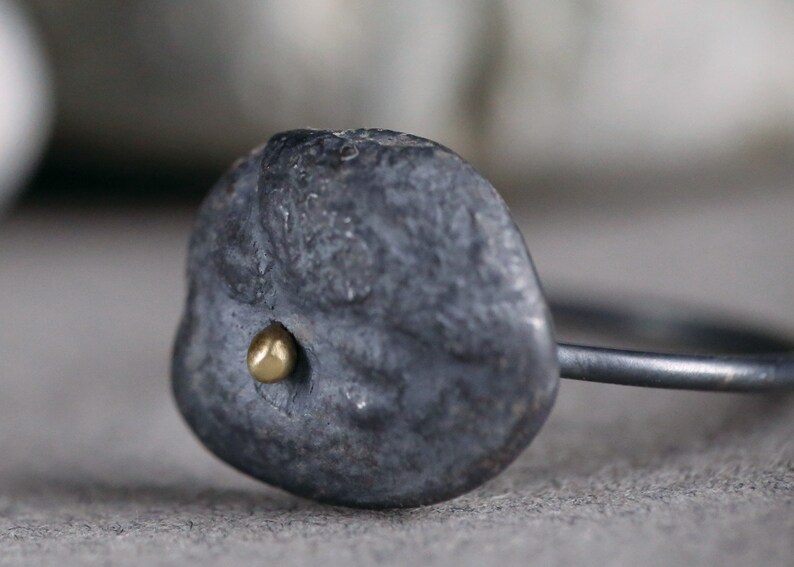 Puede incluir: Un anillo negro con una forma irregular texturizada y un peque&ntilde;o punto dorado en el centro.