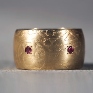 May include: A gold ring with a textured surface and two small ruby red gemstones.