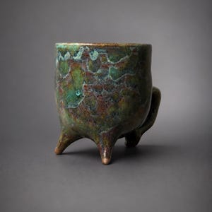 May include: A ceramic mug featuring a mottled glaze in shades of deep green, brown, and teal. The vessel has a cylindrical body supported by three short, tapered legs and includes a single curved handle attached to the side of the exterior wall.