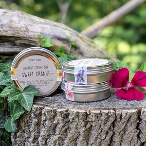 May include: Two silver tins of organic lotion bars, one labelled "Sweet Orange" with a sunflower design and the other with a lavender design. The tins are sitting on a tree stump with green leaves and a pink flower.