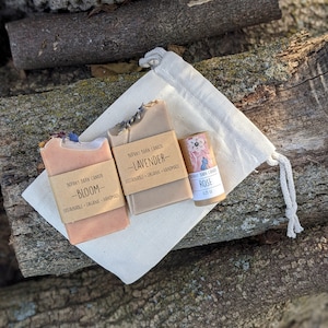 May include: Three handmade soap bars in a white muslin bag. The soap bars are labelled "Bloom", "Lavender", and "Rose". The soap bars are organic and sustainable.