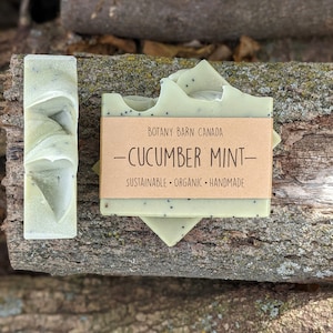 May include: Two bars of green soap with black flecks, stacked on top of each other. The top bar has a brown label that reads "Botany Barn Canada - Cucumber Mint - Sustainable. Organic. Handmade."