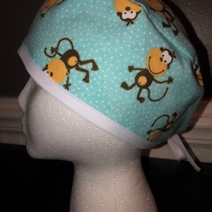 May include: A light blue scrub hat with a white trim and a pattern of brown monkeys on a white polka dot background.