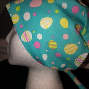 May include: A turquoise blue surgical scrub hat with a colorful Easter egg pattern. The hat has a tie that can be tied at the back of the neck.