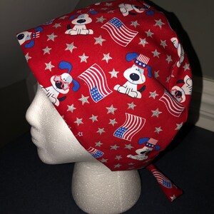 May include: Red scrub hat with a pattern of white stars, American flags, and cartoon dogs wearing red, white, and blue hats.