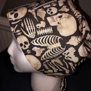 May include: A black and white skull and bones patterned surgical scrub hat. The hat is tied at the back with a long tie.