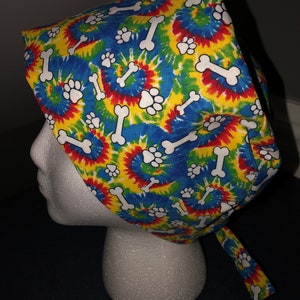 May include: A colorful tie-dye surgical scrub hat with a pattern of white paw prints and bones. The hat has a tie at the back.