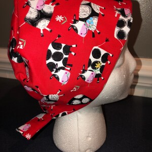 May include: A red surgical scrub cap featuring a repeating pattern of black and white cows with pink noses and floral accents. The cap is tied at the back and is displayed on a white mannequin head.