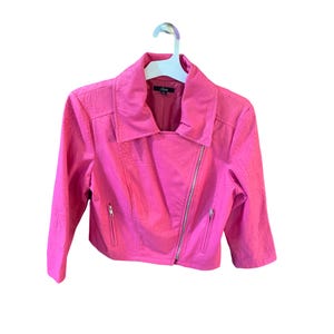 May include: A bright pink, cropped motorcycle jacket with a textured finish. The jacket features a silver zipper closure, two zippered pockets, and a notched collar. The jacket is hanging on a white hanger.