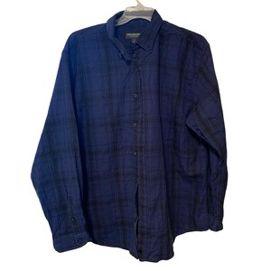 May include: A blue and black plaid button-down shirt. The long-sleeved shirt is hanging on a white hanger. The shirt has a button-down collar and a buttoned front closure. The brand name "John Bartlett" is visible on the collar.