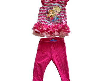 Puede incluir: Top sin mangas de rayas rosa y gris con un dobladillo con volantes y un estampado de Frozen con Elsa y Anna. El top se combina con leggings rosas.