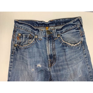 May include: A pair of blue denim jeans with distressed details and a faded look. The jeans have a classic five-pocket style and a button fly.