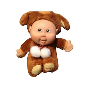 Puede incluir: Una muñeca de peluche Cabbage Patch Kid marrón que lleva un disfraz de cachorro marrón y amarillo con pompones blancos. La muñeca tiene ojos verdes y una cara sonriente.