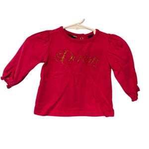 May include: A bright red long-sleeved shirt with the word "Darling" in gold script across the front. The shirt has puffed sleeves and a round neckline. The shirt is hanging on a white hanger.