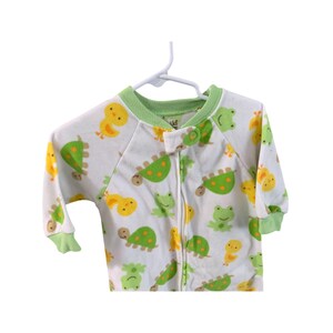 Puede incluir: Un pijama blanco para beb&eacute; con estampado verde y amarillo de tortugas, ranas y pollitos. El pijama tiene una cremallera verde y un ribete verde alrededor del cuello y las mangas.
