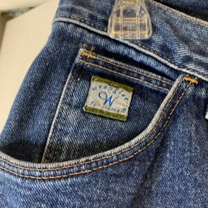 May include: Close-up of a blue denim pocket with a Wrangler for Women label sewn on. The label is green with white text.