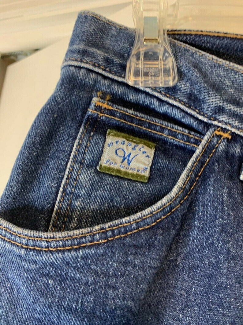 May include: Close-up of a blue denim pocket with a Wrangler for Women label sewn on. The label is green with white text.