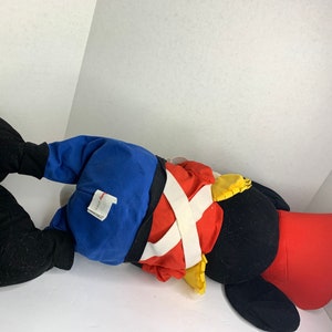 K&ouml;nnte beinhalten: Ein gro&szlig;es Pl&uuml;schtier in Form einer Zeichentrickfigur mit schwarzen, roten, blauen und gelben Farben. Das Spielzeug liegt auf der Seite und zeigt sein detailliertes Outfit mit einer roten Jacke, einer blauen Hose und einem wei&szlig;en Kreuzriemen-Design.