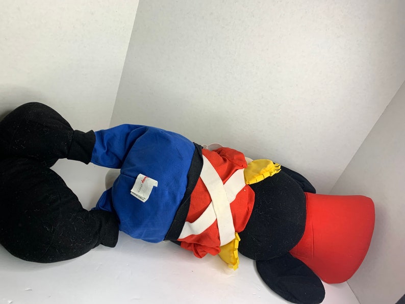 K&ouml;nnte beinhalten: Ein gro&szlig;es Pl&uuml;schtier in Form einer Zeichentrickfigur mit schwarzen, roten, blauen und gelben Farben. Das Spielzeug liegt auf der Seite und zeigt sein detailliertes Outfit mit einer roten Jacke, einer blauen Hose und einem wei&szlig;en Kreuzriemen-Design.