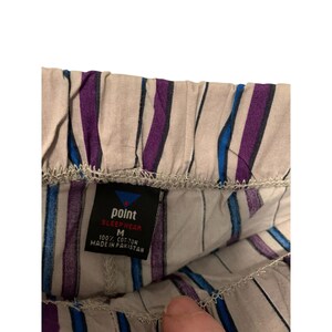 May include: Close-up of pajama bottoms with vertical stripes in purple, blue, and white. The tag reads "point sleepwear" and "100% cotton". The size is medium. The pajama bottoms are made in Pakistan.